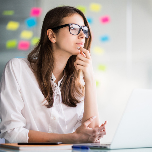Thoughtful woman wearing glasses in a white blouse at a desk, chin resting on her hand, laptop in view with colorful sticky notes on the backdrop
