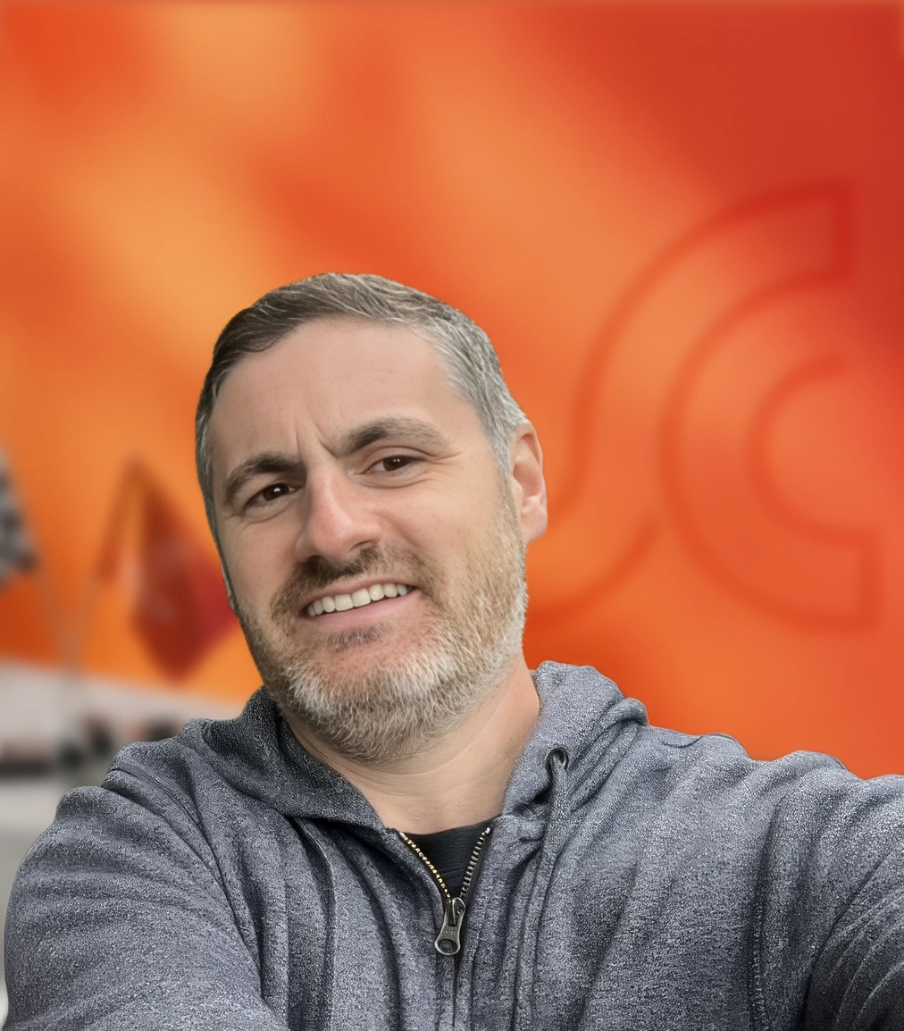 Smiling man with short gray beard taking a selfie in front of a bright orange wall, wearing a gray zip hoodie.